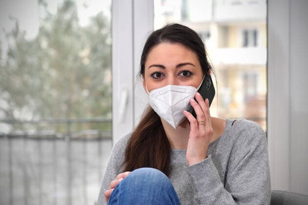 A young woman sits in her apartment working from her home office because of the Corona pandemic She is wearing an FFP2 mask and talking on a smartphone MODEL RELEASED