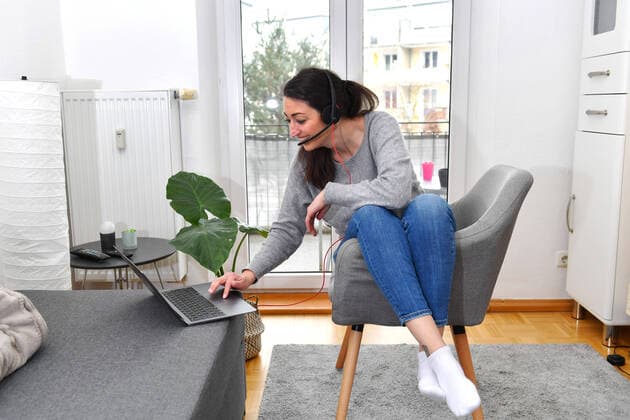 A young woman sits in her apartment with a laptop with a headset working in her home office because of the Corona Pandemic Teams, MODEL RELEASED