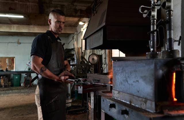 Side view of serious tattooed young male forger in apron heating metal in furnace while working in grungy smithy Copyright: xDanielxDexAndresx Model Released