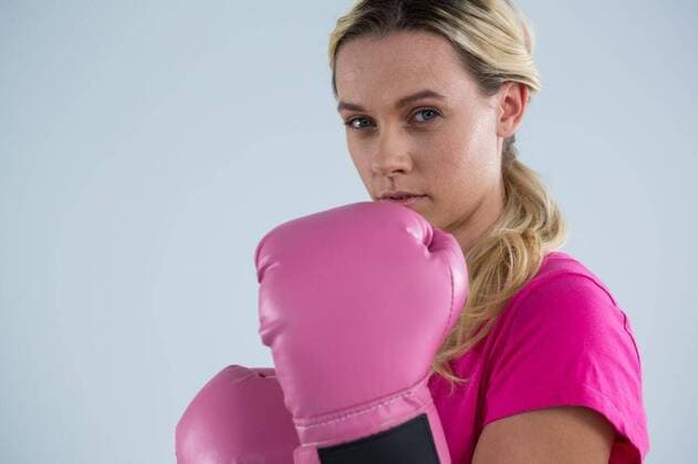 IMAGO Creative: Brustkrebsmonat Portrait of young woman with boxing gloves Portrait of young woman with boxing gloves standing against gray backgrond, model released, Cape Town, South Africa, 26.04.2017, 1055134.jpg, gray background, casual clothing, cut out, breast cancer, pink, awareness, womens health, cancer, illness, breast cancer awareness, healthcare and medicine, disease, moral support, support, tumor, fight against cancer, millennial, lifestyle, standing, blond hair, fighting, facial expression, serious, gloves, boxing, determination 20s, Young Adult, Woman, Female, Caucasian, 25-29 Years, Young Women, Caucasian Ethnicity Studio shot,Side view,Portrait,Looking at camera,Head and shoulders