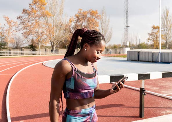 IMAGO Creative: Sport Start-Up Smiling sportswoman with braided hair using smart phone on track during sunny day model released Symbolfoto JCCMF00888