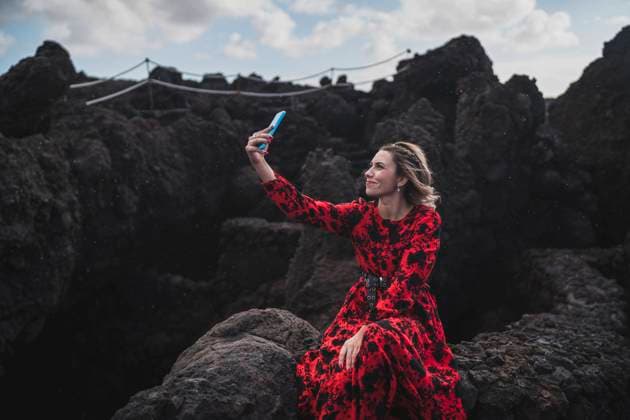 IMAGO Creative: Welttourismustag am 27. September Young woman smiling while taking selfie through mobile phone sitting on rock at Los Hervideros, Lanzarote, Spain model released Symbolfoto SNF00969