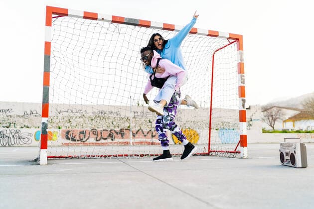 IMAGO Creative, Urban Lifestyle IMAGO Creative: Urban Diversity Cheerful woman showing horn gesture while riding piggyback on African American boyfriend on sports ground with retro tape recorder Copyright: xLuisxManuelxMunozx Model Released