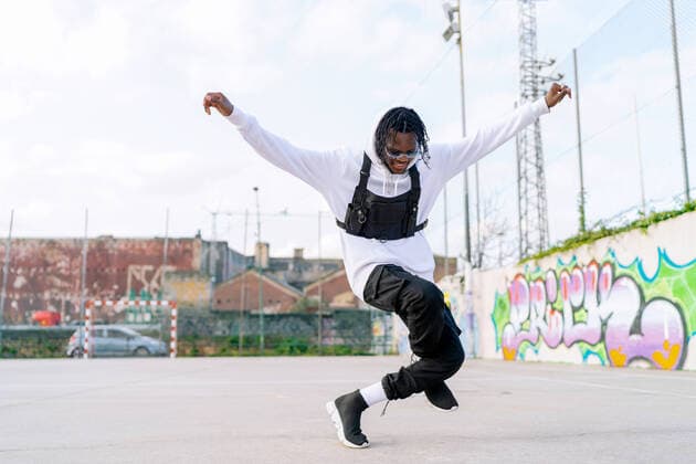 Young trendy ethnic man in sunglasses with stretched arms dancing on tiptoes while looking down against graffiti wall in city Copyright: xLuisxManuelxMunozx Model Released