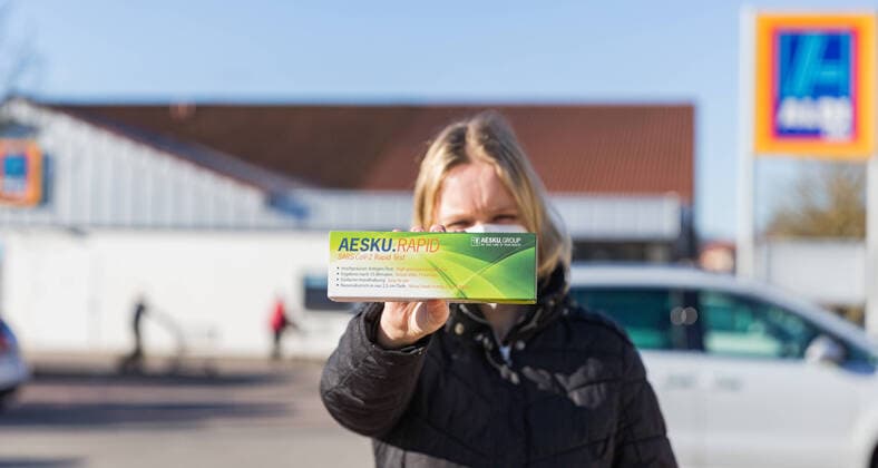 06 03 2021, Corona Rapid Tests at Aldi Available from today Already today, Saturday, ALDI Nord and ALDI Süd offer so-called Corona Antigen Rapid Tests Self-tests for sale Drugstores like Rossmann and Co follow suit on Monday A woman stands in front of an ALDI Süd store holding an AESKU RAPID SARS CoV 2 Rapid Test Bad Brueckenau Aldi Süd Bava
