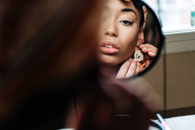 IMAGO Creative: Clickbait, Hautpflege und Kosmetik Reflection of content African American female putting on earring and looking in round mirror at home Copyright: xJakexJakabx Model Released