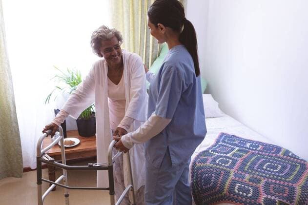 IMAGO Creative: NGO, Rehabilitation und Gesundheitswesen Female nurse helping senior female patient to stand with walker Female nurse helping senior female patient to stand with walker at nursing home Cork, Ireland, 26.02.2019, 1242845.jpg, senior, retirement, pensioner, uniform, nurse, nursing, expertise, occupations, patient, shelter, daycare, accommodation, nursing home, retirement home, clinic, retiree, old-age, senior citizen, active, active senior, bed, blanket, standing, talking, interacting, holding, holding hand, helping, walking, disabled, handicapped, physical impairment, walker, health care, medical examination, checkup, medicine, treatment, together, care, trust, lifestyle, leisure Indoors,Front view,Half length