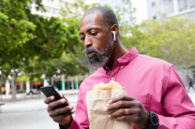 African American man using his phone and eating a takeaway sandwich African American man out and about in the city streets during the day, wearing pink shirt and earphones, having a takeaway snack and using a smartphone., model released, , property released, Cape Town, South Africa, 11.03.2020, 1334722.jpg, people,real people,commuting,commuter,travel,city,creative,individuality,lifestyle,street fashion,modern,on the go,digital nomad,independent,casual,street,smartphone,mobile phone,technology,phone,holding,using,communication,connection,interacting,earphones,listening,musing,smartwatch,takeaway,to go,snack,lunch,street food,eating,paper bag Mature Adult,Man,Male,Black Outdoors,Side view,Day,Half length