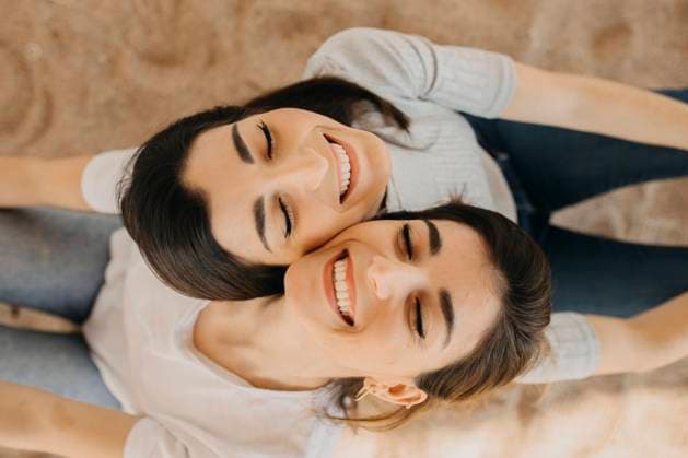 IMAGO Creative: Schwesternschaft Smiling sisters with eyes closed on sand at beach model released Symbolfoto ARTF00041
