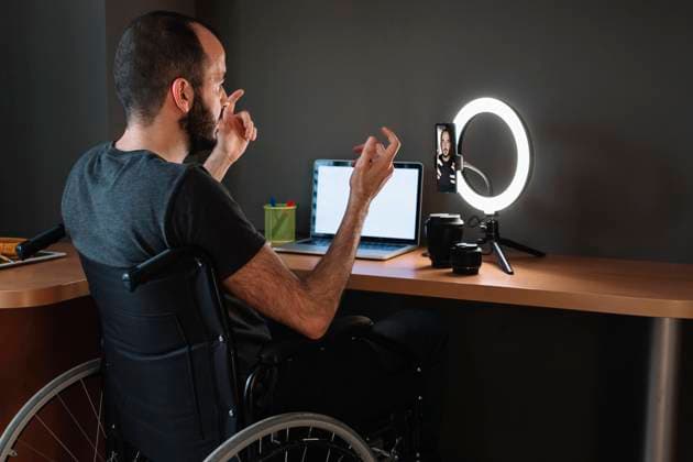 Vlogger filming through mobile phone with circular strobe tripod while sitting on wheelchair at studio model released Symbolfoto property released XLGF01348