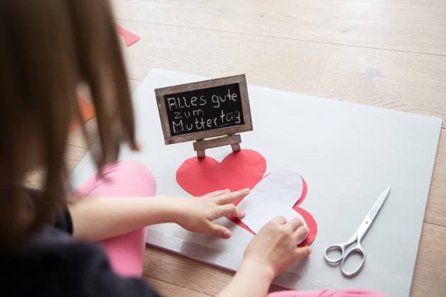 Bamberg, Deutschland 04. Mai 2021: Ein kleines Mädchen sitzt auf einem Laminatfussboden und bastelt zum Muttertag für die Mutter ein Herz aus Pappe als Geschenk, daneben liegt eine Schere. Auf einer Tafel steht mit Kreide geschrieben, alles gute zum Muttertag. Bayern *** Bamberg, Germany 04 May 2021 A little girl sits on a laminate floor and makes a cardboard heart as a gift for her mother for Mothers Day, next to it lies a pair of scissors On a blackboard is written with chalk, Happy Mothers Day Bavaria Copyright: xFotostandx xK.xSchmittx