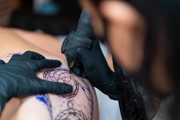 IMAGO Creative: Neue Aussichten Female tattooer in cloth face mask with machine drawing tattoo on body of unrecognizable client in salon Copyright: xEnriquexEspinosax Model Released