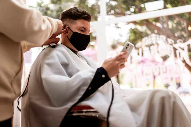 From below of crop stylist with trimmer cutting hair of man in cloth face mask with cellphone in barbershop Copyright: xJavierxBallesterx Model Released