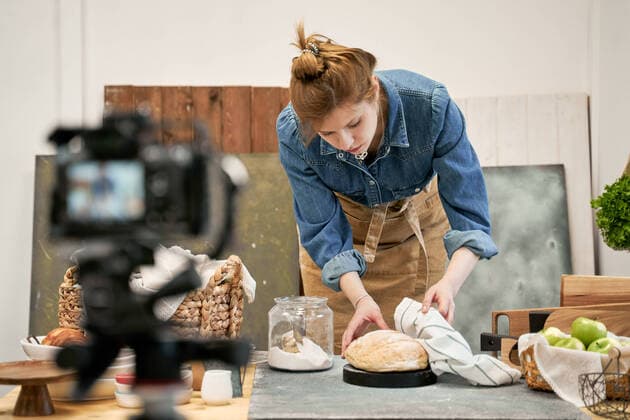 Young female blogger with fresh wheat bread against photo camera during cooking process at table in house, Model released Copyright: xSergeyxMironovx MR