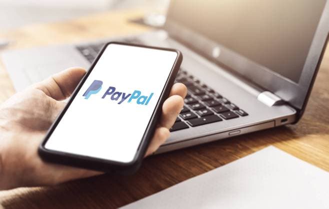 Bavaria, Germany - 18 May 2021: Paypal online payment service and banking company on a smartphone display that is held in the hand in front of a notebook on a table. Paypal online payment