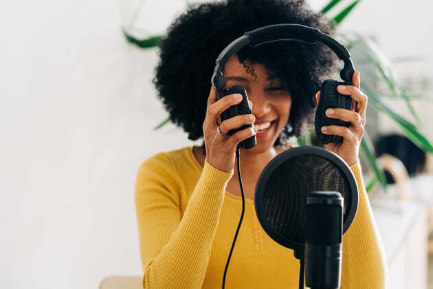 Cheerful young black female with Afro hair in casual clothes and headphones smiling and recording podcast sitting at table with laptop and microphone in light studio Copyright: xJakexJakabx Model Released