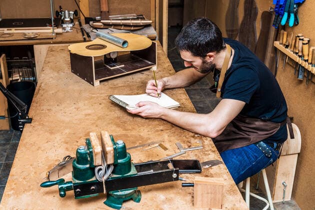 Side view of adult male artisan taking notes in notebook at table with guitar sound board in workroom Copyright: xLuisxManuelxMunozx Model Released
