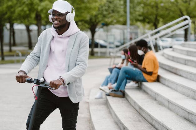 African American male in wireless headphones riding electric scooter with unrecognizable friends sitting on stairway on the background, Model released Copyright: xOierxAsox MR