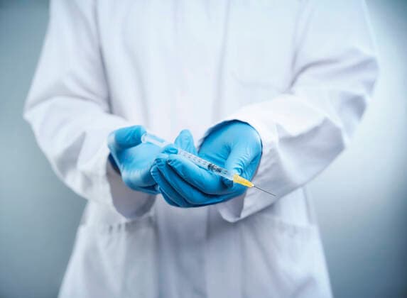 Female doctor wearing lab coat and protective glove holding COVID-19 vaccine syringe model released Symbolfoto DIKF00589