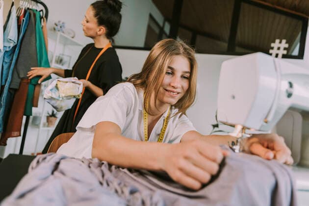 Smiling teenage fashion designer sewing cloths with mature woman in background working at workshop model released Symbolfoto property released MFF08166
