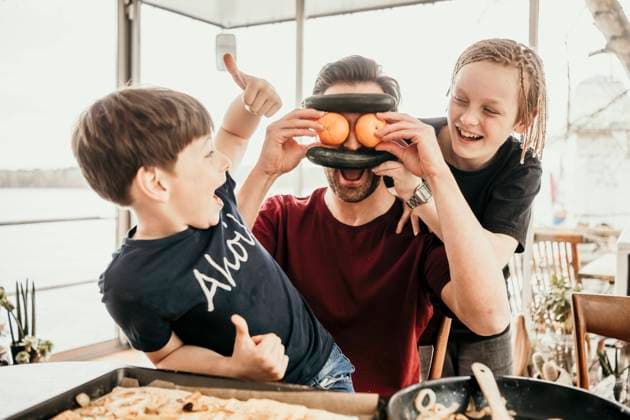 Cheerful siblings looking at playful father covering face with fruits and vegetables in kitchen at houseboat model released Symbolfoto property released MJRF00577