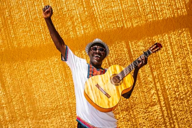 Cheerful man with hand raised carrying guitar in front of yellow wall during sunny day model released Symbolfoto OCMF02148