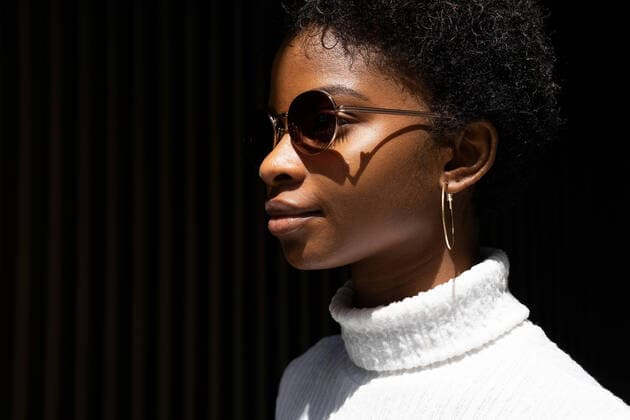 Creative Highlights Symbolbilder Side view of young African American woman in stylish sweater and sunglasses looking away while standing in bright sunlight against black background Copyright: xDavidxMunozx Model Released