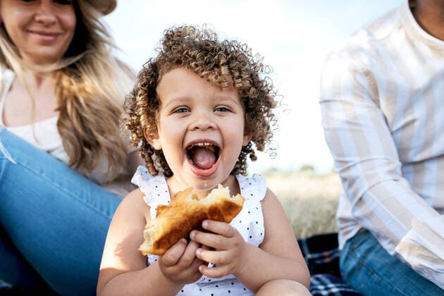 Creative Highlights Symbolbilder Cheerful little girl eating pastry looking at camera sitting with multiracial family enjoying picnic together in nature Copyright: xSergioxVictorxVegax Model Released