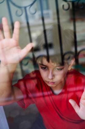 Through glass of unhappy preteen boy with bruises on face looking away while standing near window at home as concept of domestic violence and child abuse, Model released AdalbertoRodriguez_ChildAbuse_46.jpg Copyright: xAdalbertoxRodriguezx MR