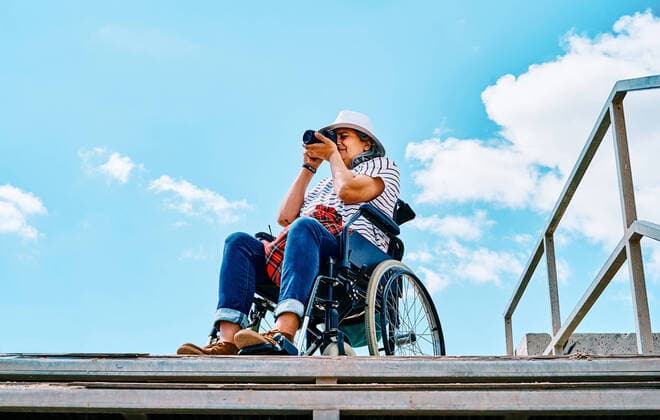 From below gray haired disabled woman in wheelchair taking pictures on photo camera near stairway against blue sky in city, Model released AdalbertoRodriguez_WomanInWheelchairBeach_25.jpg Copyright: xAdalbertoxRodriguezx MR