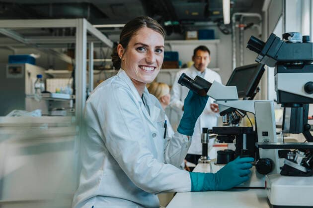Smiling woman analyzing human brain slide under microscope while sitting with scientists in background at laboratory model released Symbolfoto property released MFF06495