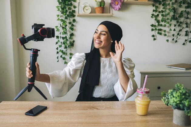 Smiling female vlogger waving while filming through camera at home model released Symbolfoto property released JCCMF03472