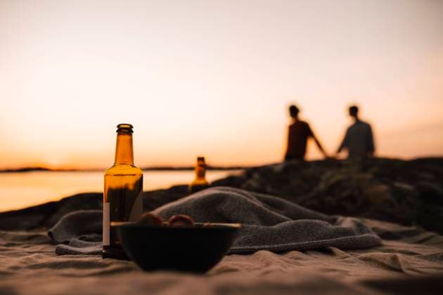 Beer bottle and bowl on picnic blanket while gay couple in background at lakeshore during sunset Sweden, Gothenburg, model released Copyright: xMaskotx , MA88016