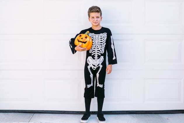 Full body of smiling preteen boy wearing black Halloween costume with skeleton print standing near carved Jack O Lantern pumpkin against white wall, Model released DavidBenito_HalloweenKids_6.jpg Copyright: xDavidxBenitox MR