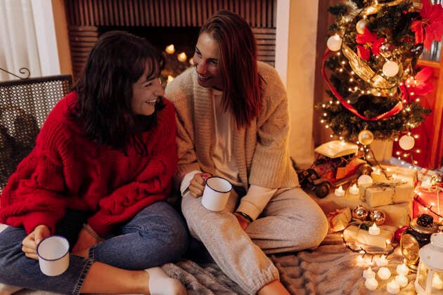 Smiling female friends with cup sitting by Christmas tree at home model released Symbolfoto property released EGHF00174
