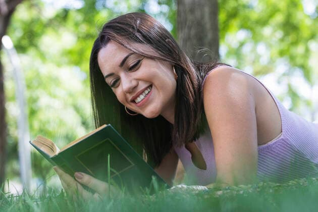 Smiling young woman reading book while lying on grass in park model released Symbolfoto PNAF02162