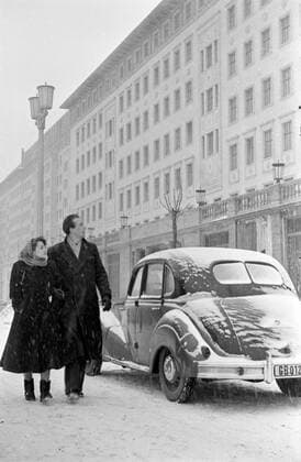 Stalinallee im Schnee Winter Berlin 1951 Block C Südseite Historisches Bild und Foto: Ehepaar läuft im Winter bei Schneeregen an den Neubauten der Stalinallee, Block C entlang. Auto im Hintergrund. Stalinallee Berlin Winter 1951 Block C Südseite. Bau des Block C Südseite, Portal und Gebäude, Architekt Richard Paulick der Stalinallee. Bau, Umbau der Stalinallee im Jahr 1951 nach den Visionen des Sozialistischen Realismus bzw. der Nationalen Bautradition. VEB Industriebau. Errichtung der Arbeiterpalaeste. Die Strasse, boulevard wurde zwischen 1949 und 1961 Stalinallee (Große Frankfurter Straße) genannt und war das Vorzeigeprojekt im Rahmen des Ostdeutschen Wiederaufbauprogamms nach dem zweiten Weltkrieg. Die Stalinallee wurde in mehrer