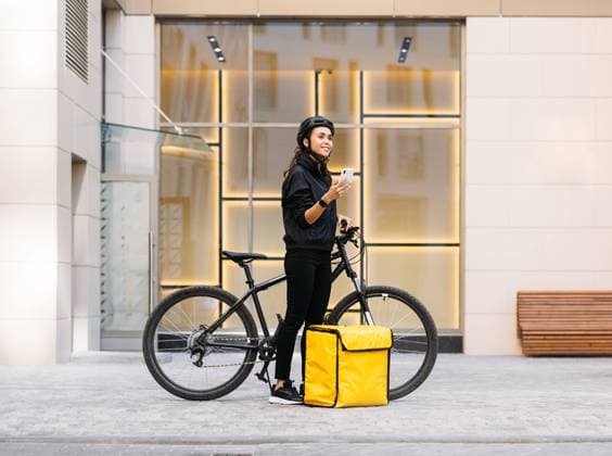 IMAGO Creative: 2021 Trends Side view of woman in uniform with bicycle and food delivery backpack holding a mobile phone while standing at apartment building Copyright: xxArtemxVarnitsinx