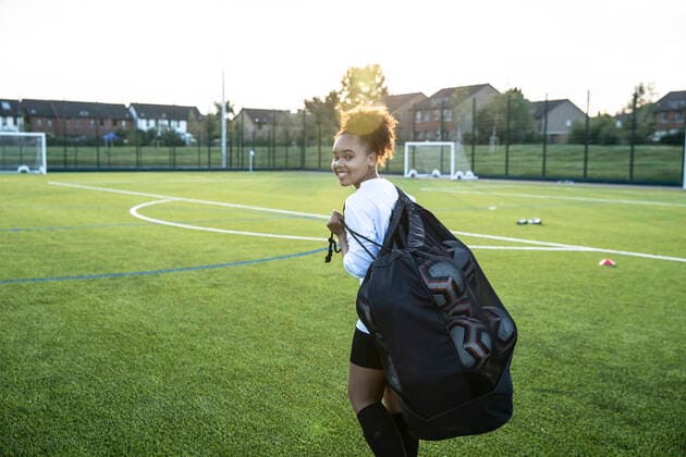 UK, Smiling female soccer player (12-13) carrying bag with balls in field UK, Smiling female soccer player carrying bag with balls in field