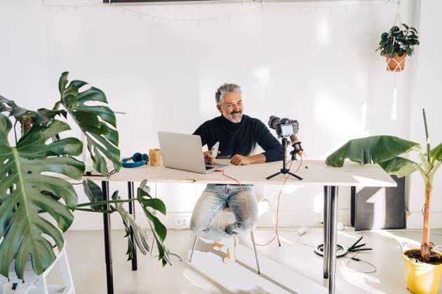 Smiling aged broadcaster sitting at table with laptop near potted plants and looking at camera recording while creating video podcast JakeJakab_PedroAtLoft_40.jpg Copyright: xJakexJakabx Model Released