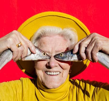 Positive aged female with gray hair in stylish yellow hat covering eyes with raw fish while standing on red background, Model released SergioVictorVega_TitiFishes_1.jpg Copyright: xSergioxVictorxVegax MR