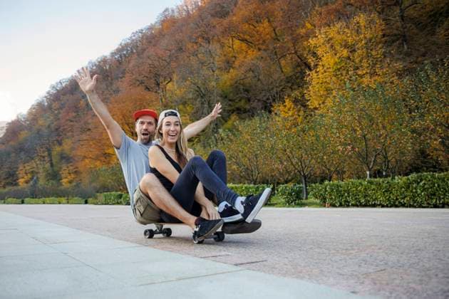 IMAGO Creative: Fit durch den Herbst Man with arms raised sitting behind woman on skateboard at footpath model released, OMIF00247