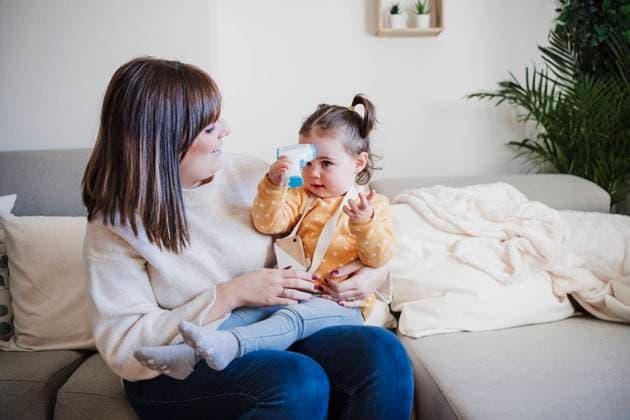 Smiling mother looking at cute baby girl holding infrared thermometer on sofa model released property released, EBBF05093