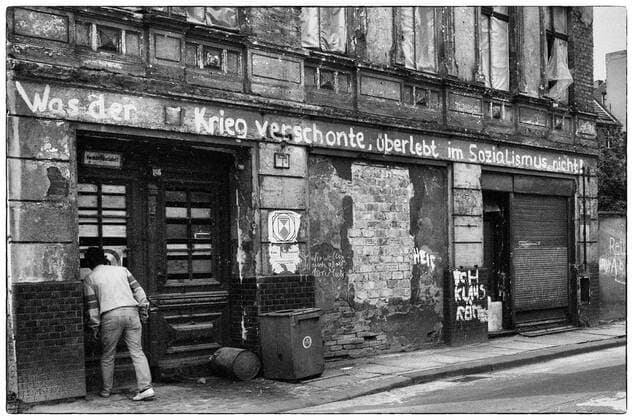 02.04.1990, Berlin, DDR - Spruch an der Fassade eines Wohnhauses: Was der Krieg verschonte, überlebt im Sozialismus nicht!. (20-30 Jahre, Alltag, aussen, Aussenaufnahme, Berlin, DDR, deutsch, Deutsche Demokratische Republik, Deutschland, Europa, europaeisch, Fassade, Fruehjahr, Fruehling, GDR, Gebaeude, Gesellschaft, Haus, Hausfassade, historisch, Jahreszeit, Kaukasier, kaukasisch, Mangelwirtschaft, Mann, marode, Mensch, Misswirtschaft, Ostberlin, Person, QF, Querformat, restaurierungsbeduerftig, ruinoes, s w, sanierungsbeduerftig, schwarz-weiss, Schwarzweiss, Spruch, Stadt, Stadtbild, Stadtlandschaft, Strasse, Strassenszene, Verfall, verfallen, Was der Krieg verschonte ueberlebt im Sozialismus nicht, Westeuropa, Wirtschaft, Wohngebaeude, W