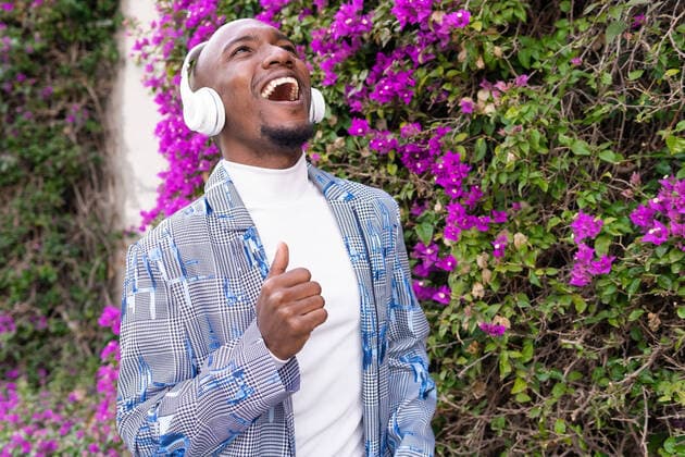 Side view of delighted African American male in stylish outfit looking up listening to music in wireless headphones while standing near bush with blooming flowers on summer day, Model released DavidMunoz_MarcBcn_69.jpg Copyright: xDavidxMunozx MR