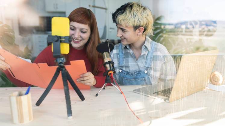 Cheerful female friends sitting at table with documents and microphone while shooting podcast on smartphone in light room at home JoseCarlosIchiro_CoupleWomaenAtLoft_02.jpg Copyright: xJosexCarlosxIchirox Model Released