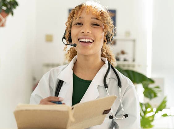 IMAGO Creative: Lokalhelden - Gesundheits- und Rettungskr?fte Smiling ethnic female doctor in headset with mic and stethoscope on neck and notebook preparing for video call, Model released, Property released JoseCarlosIchiro_WomenAtHome_50.jpg Copyright: xJosexCarlosxIchirox MR