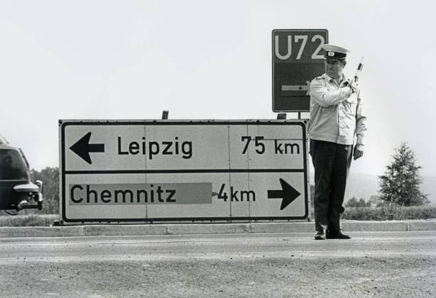 DDR Verkehrspolizist regelt den Verkehr bei Chemnitz, Juli 1990 DDR, bei Chemnitz, Juli 1990, Verkehrspolizist, DDR-Verkehrspolizist, regelt Verkehr mit Verkehrsregelstab, Naehe Chemnitz, Richtungsschild Karl-Marx-Stadt mit dem neuen alten Städtenamen Chemnitz überklebt, Straße zwischen Chemnitz und Leipzig, Gestik, Volkspolizei, Vopo, Uniform, Verkehr, Strassenverkehr, regeln, regelt, Verkehrsregelung, Verkehrsvorschriften, Deutsche, DDR-Polizei, Verkehrspolizei, DDR-Verkehrspolizei, Polizist, DDR-Polizist, Polizeiuniform, Polizei, Polizeistab, Reglerstab, Reglerstaebe, Stab, Verkehrsstab, Verkehrsregeln, Ostalgie, DDR, DDR-Verkehr, Geschichte, Wende, Wendezeit, 1990er, 90er Jahre, Neunziger Jahre, Zeitgeschichte, historisch, *** DDR tr