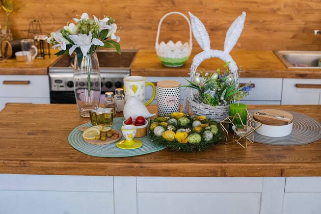 Beautiful spring Easter kitchen counter. flowers and decorative eggs. Vase of lilies on the counter. Still life of painted Easter eggs in wreath with lemon and olives and tomatoes on wooden kitchen counter. A lovely basket with bunny ears with flowers and decorative eggs. Vase of lilies on the counter. Copyright: xx 42724050