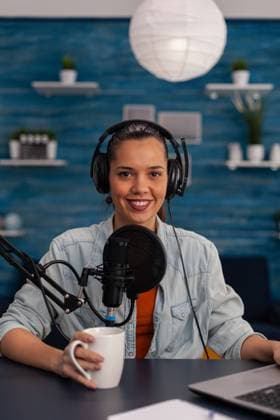 Portait of woman vlogger holding cup and streaming podcast using laptop. Portait of woman vlogger holding cup and streaming podcast using laptop. Social media influencer recording livestream with headphones and microphone in home office. Smiling blogger podcasting. Copyright: xPhotographer:xDragosxCondreax 43201956